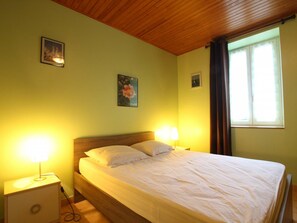 A bedroom with a wooden ceiling, a bed with white bedding, two bedside lamps, and a window with curtains. - Charming Beachside Home with Terrace, BBQ, and Baby Equipment in Hauteville-sur-Mer (Hauteville-sur-Mer)
