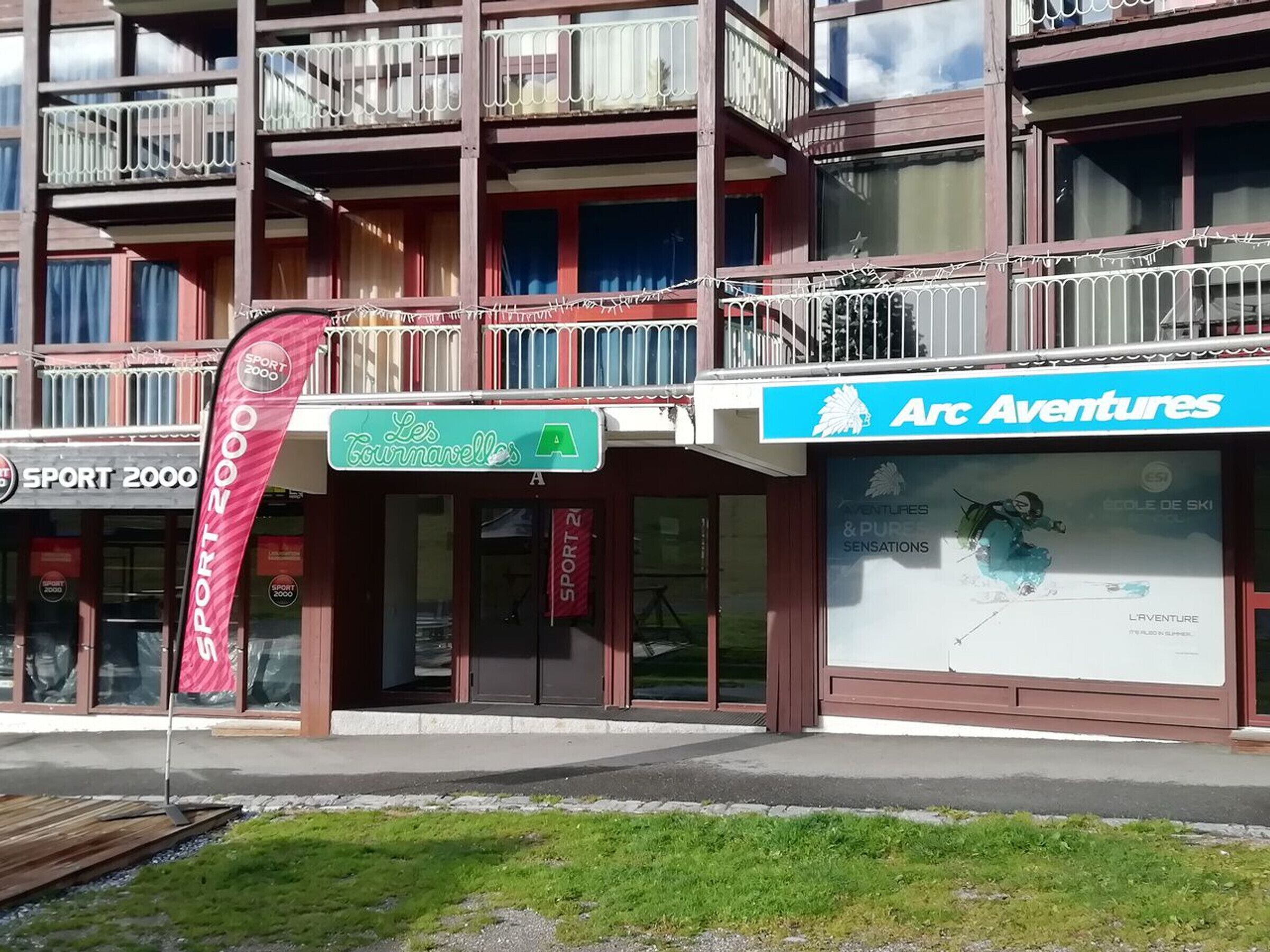 Un edificio a più piani con balconi, una vetrina con il cartello 'Arc Adventures' e una bandiera rossa con il logo 'SPORT 2000'.
