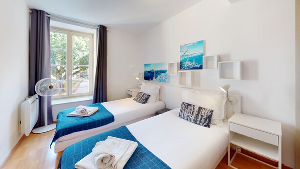 Een hotelkamer met twee bedden, een raam met gordijnen, een ventilator aan de muur en kunstwerken.