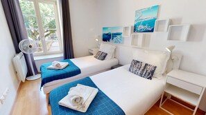 A hotel room with two beds, a standing fan, a window with curtains, and artwork on the wall.