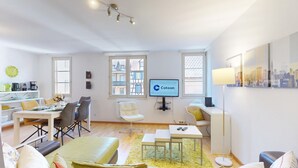 Een moderne woonkamer met een eethoek, een aan de muur bevestigde televisie en een stadslandschap schilderij.