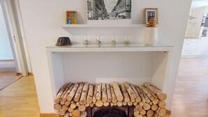 Een kamer met een tafel met glazen blad waarop een foto uit vervlogen tijden, een klein beeldje en drie glazen potten staan. Onder de tafel staat een bank met keurig opgestapeld hout.