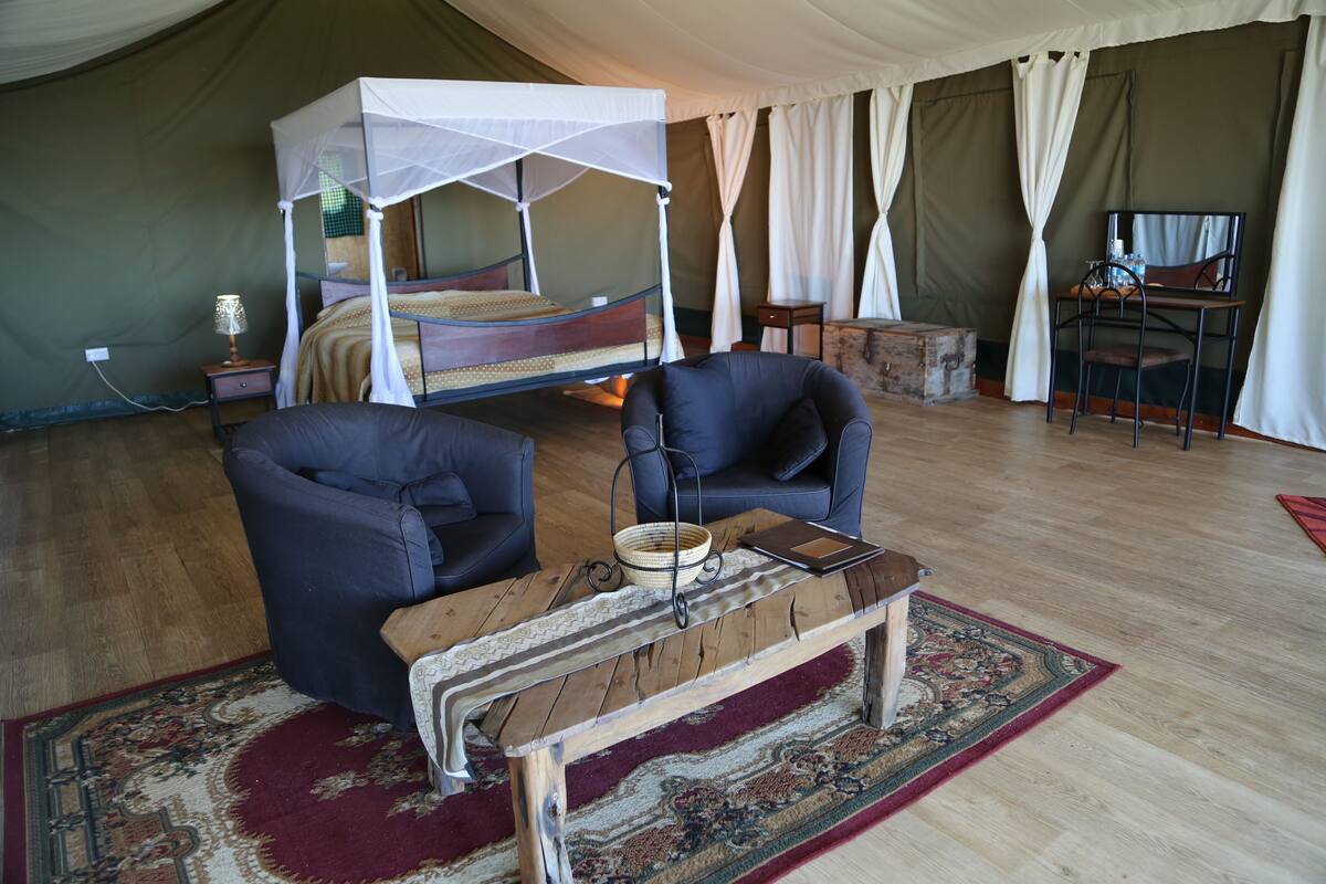 A tented accommodation room with a bed, two chairs, a table, and a small desk with a chair.