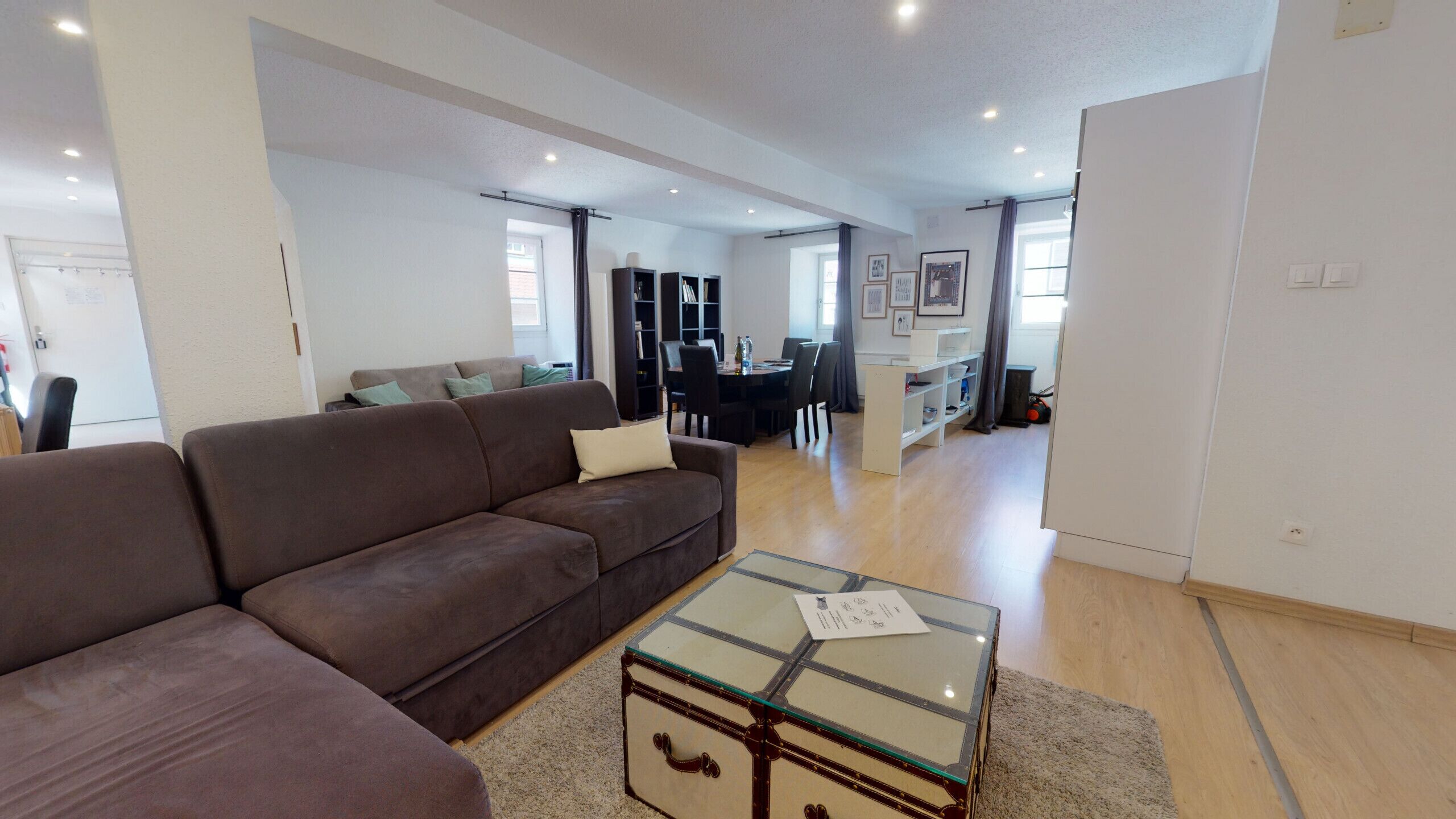 A modern living room with a brown sofa, a glass-top coffee table, and a dining area in the background.