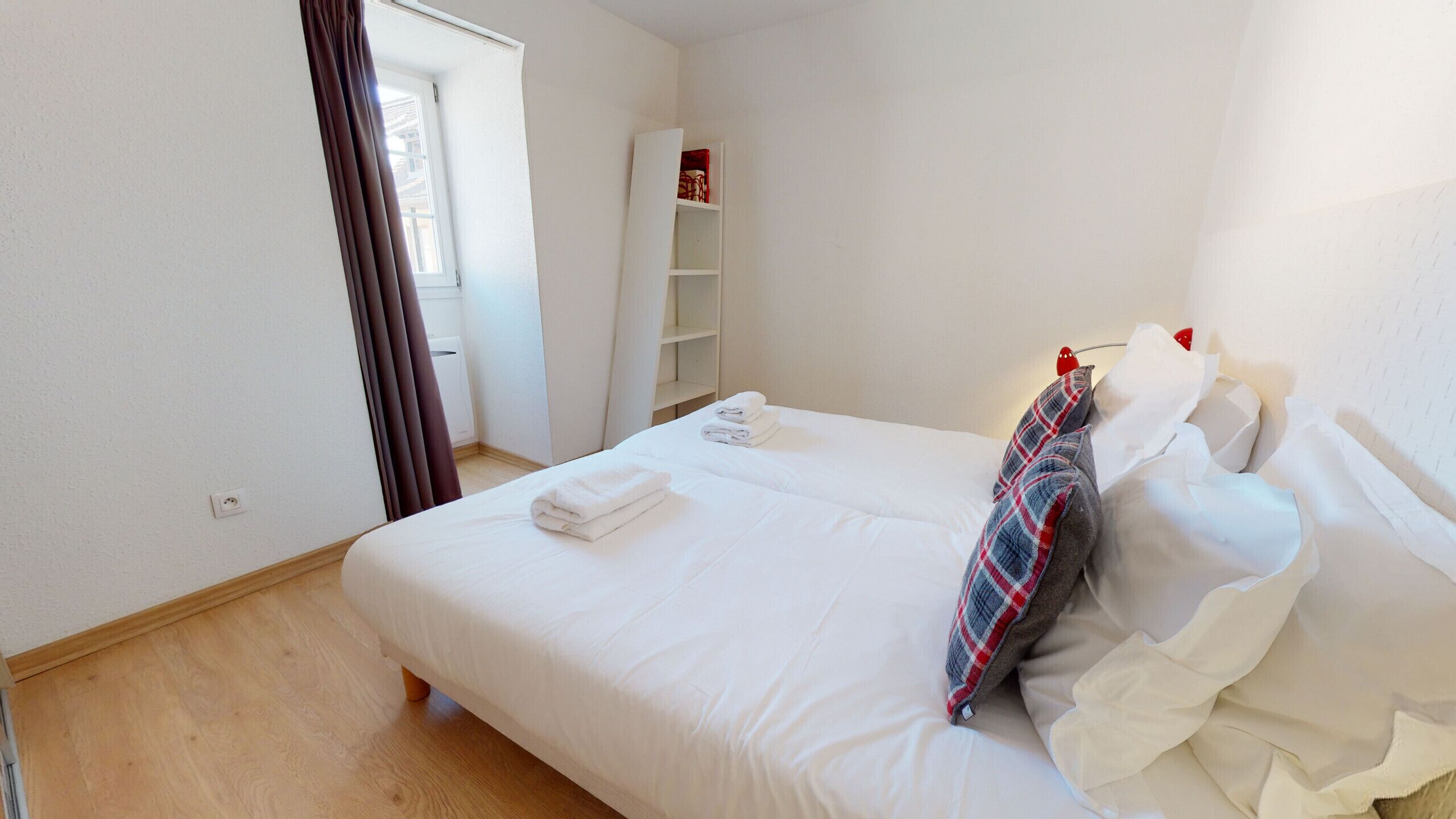 A neatly made bed with white linens and a plaid pillow, a white wardrobe, and a window with curtains.