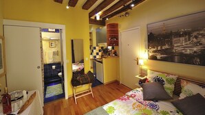 A room with a bed, a desk, a chair, a small table with a bottle, a handbag, and a painting of a cityscape. - A1DG Home Sweet home (París)