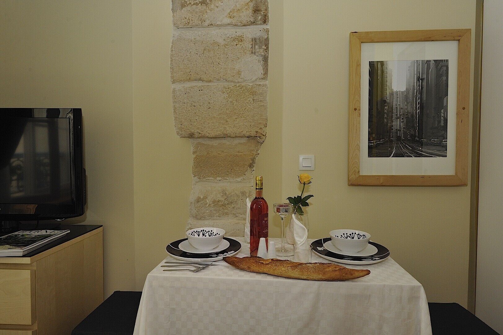 A dining table with a white tablecloth, two white bowls with black designs, a bottle of red wine, a glass of water, a vase with a yellow flower, and a long bread roll. A framed black and white photo hangs on the wall above the table.