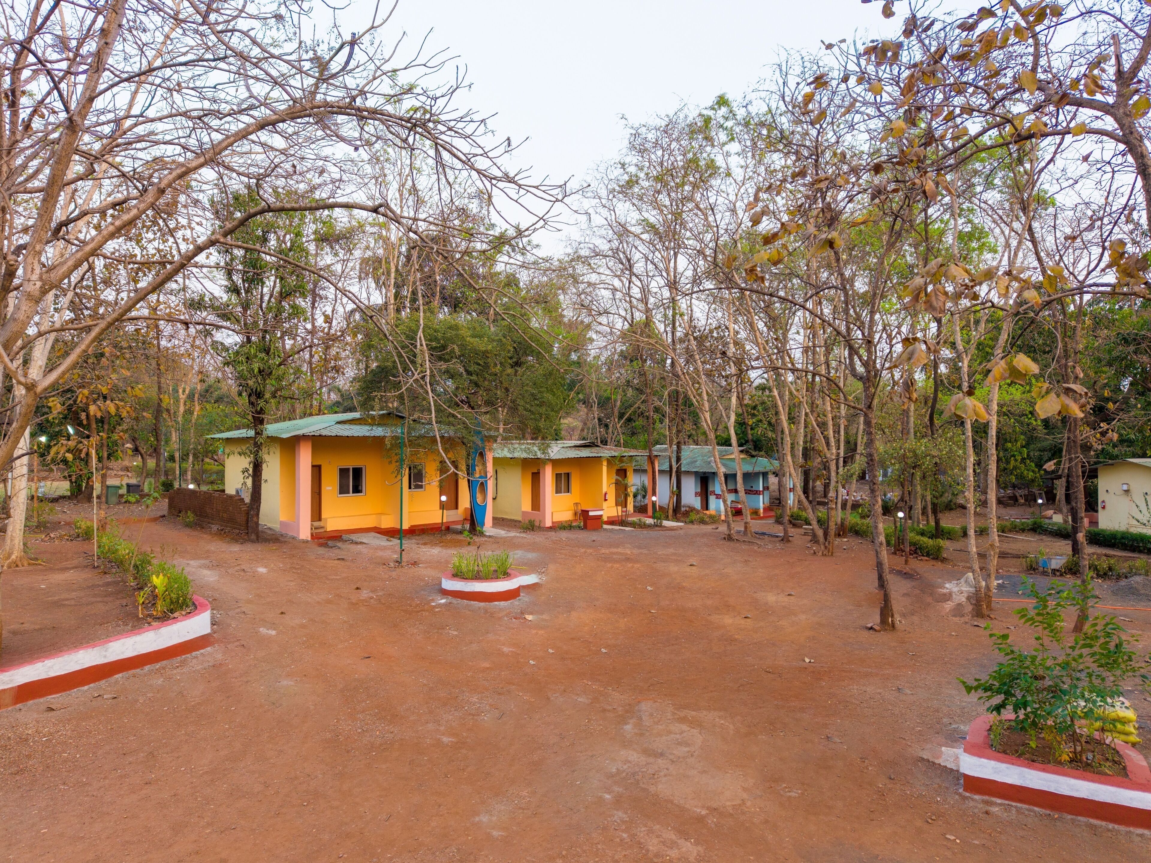 A rustic outdoor area with multiple small, single-story buildings, surrounded by trees and a dirt ground.