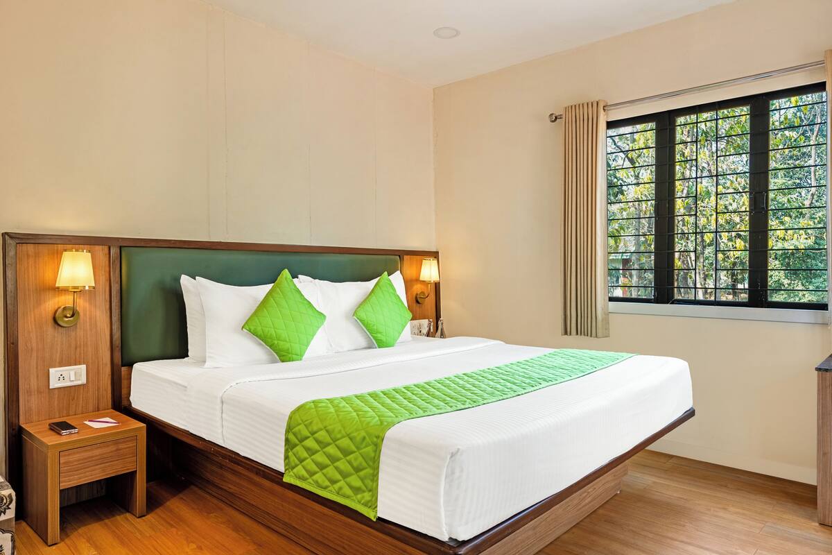 Quarto de hotel com uma cama grande, duas luminárias de cabeceira, mesa de apoio, janela com vista para árvores e piso de madeira.