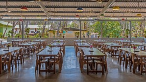 A covered outdoor dining area with multiple tables and chairs, a central pathway, and a view of trees and buildings in the background. - Nature Trails Sajan, Vikramgad (Vikramgadh)