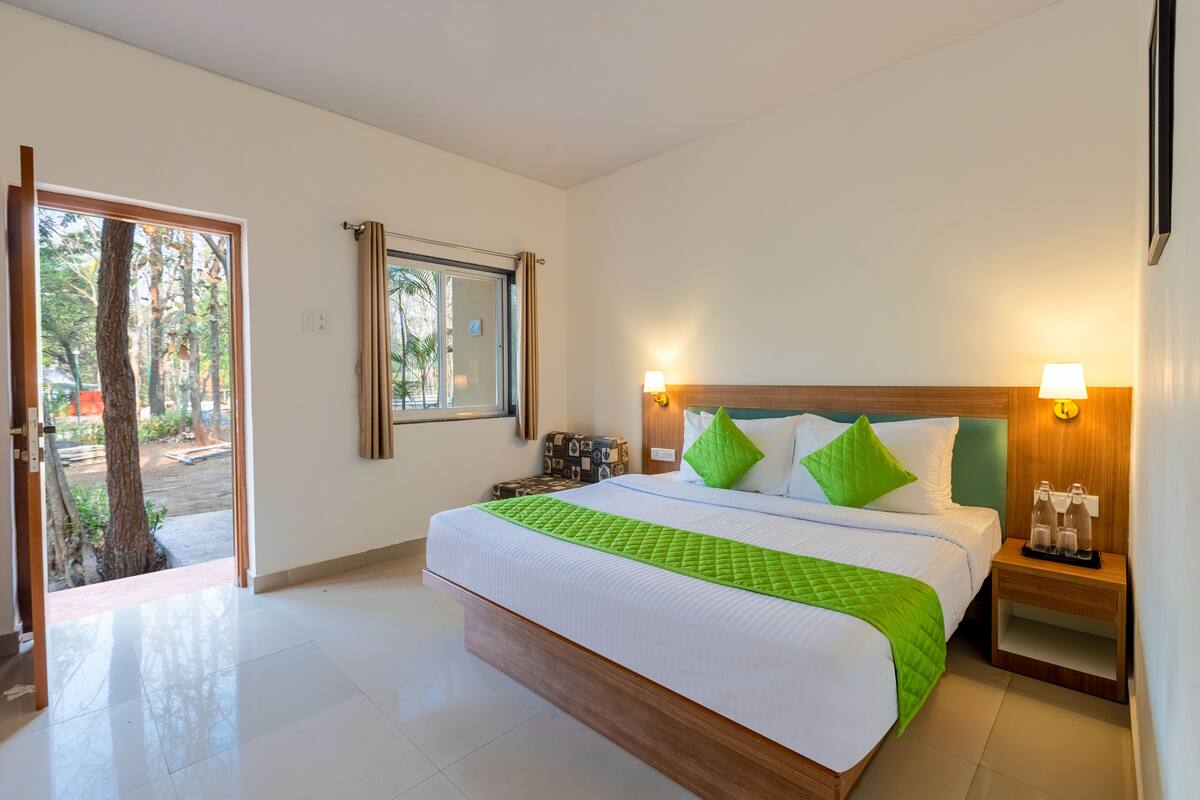 Quarto de hotel com uma cama grande, cabeceira de madeira, duas luminárias de cabeceira, mesa de apoio e vista para o exterior.