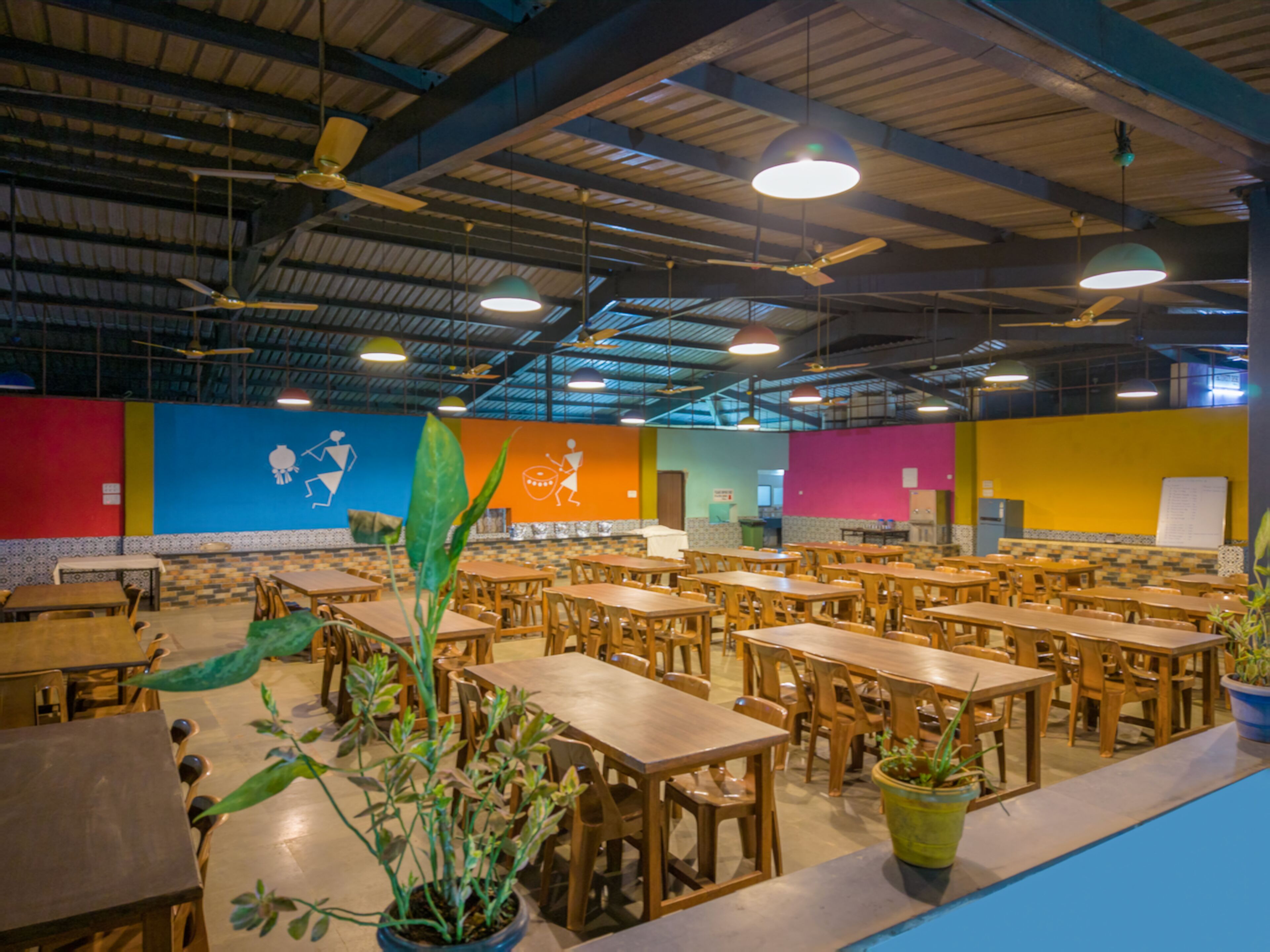 Un amplio comedor interior con varias mesas y sillas de madera, ventiladores de techo y plantas en macetas.