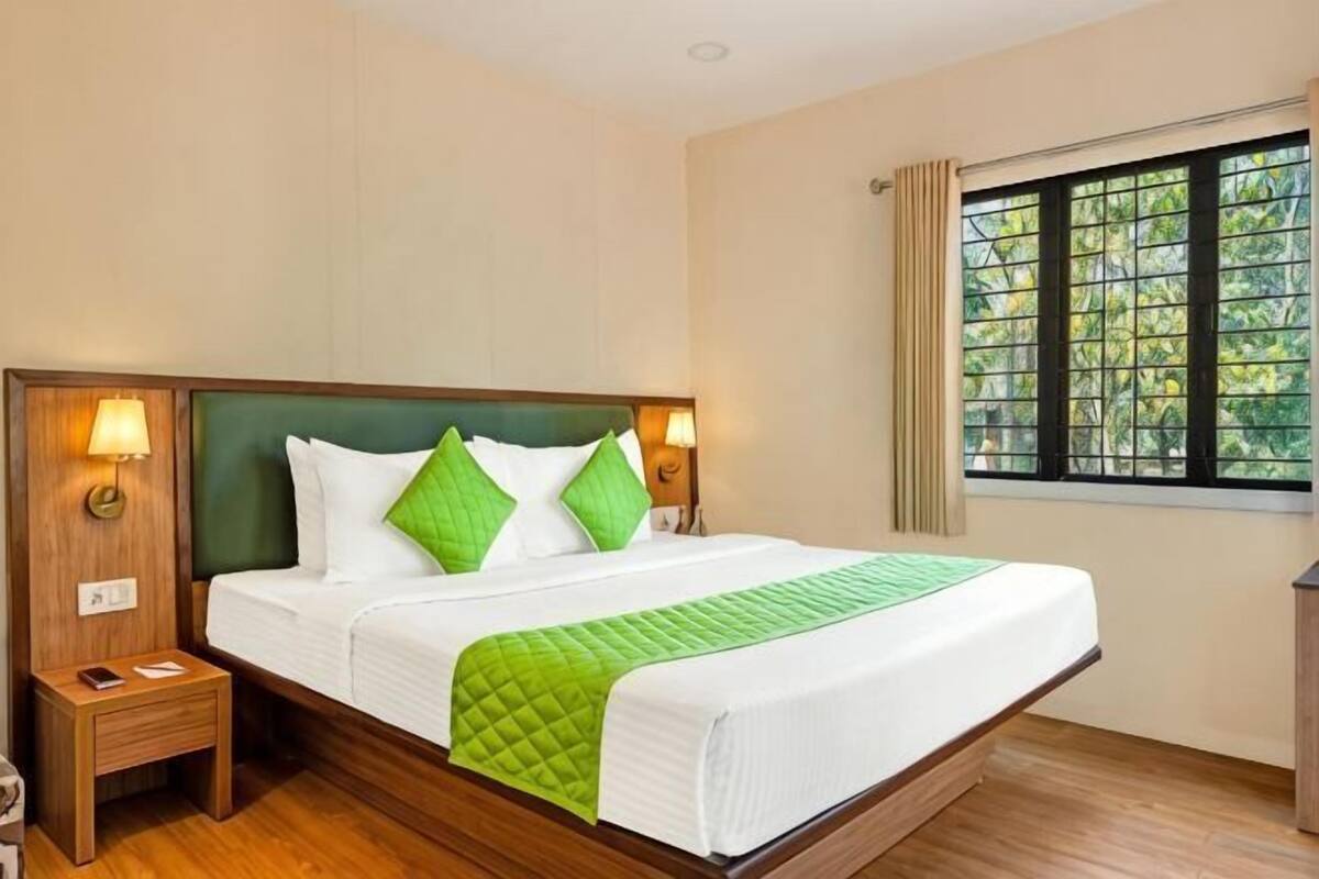 Quarto de hotel com uma cama grande, duas luminárias de cabeceira, um criado-mudo e uma janela com vista para árvores.