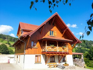 A wooden house with a sloped roof, a balcony with flower pots, and a stone wall in the foreground.
