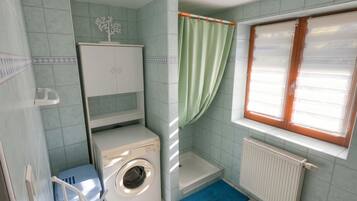 A compact bathroom with a washing machine, a radiator, and a window with curtains.