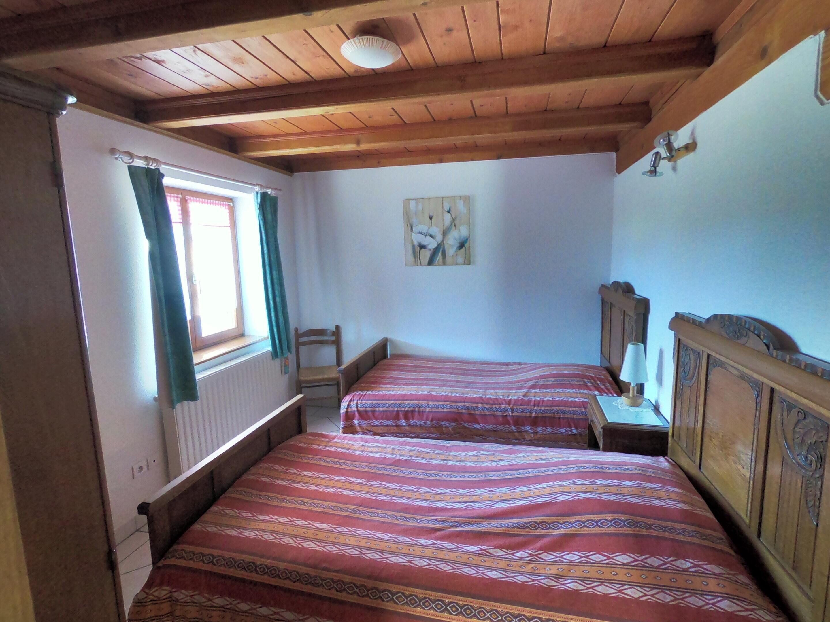 A bedroom with two wooden beds, a window with curtains, a radiator, and a painting on the wall.