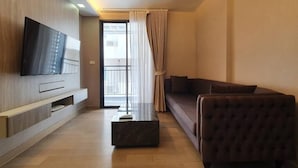 A modern living room with a flat-screen TV mounted on the wall, a sofa, a coffee table, and a sliding glass door leading to a balcony. - Dazzle Residence (Bangkok)