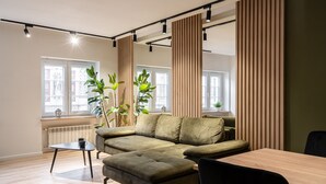 Una sala moderna con un sofá modular, una mesa de comedor y una mesita auxiliar. Hay ventanas amplias con cortinas, un radiador y plantas en macetas.
