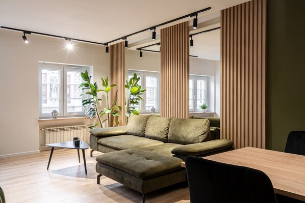 Una sala moderna con un sofá modular, una mesa de comedor y una mesita auxiliar. Hay ventanas amplias con cortinas, un radiador y plantas en macetas.