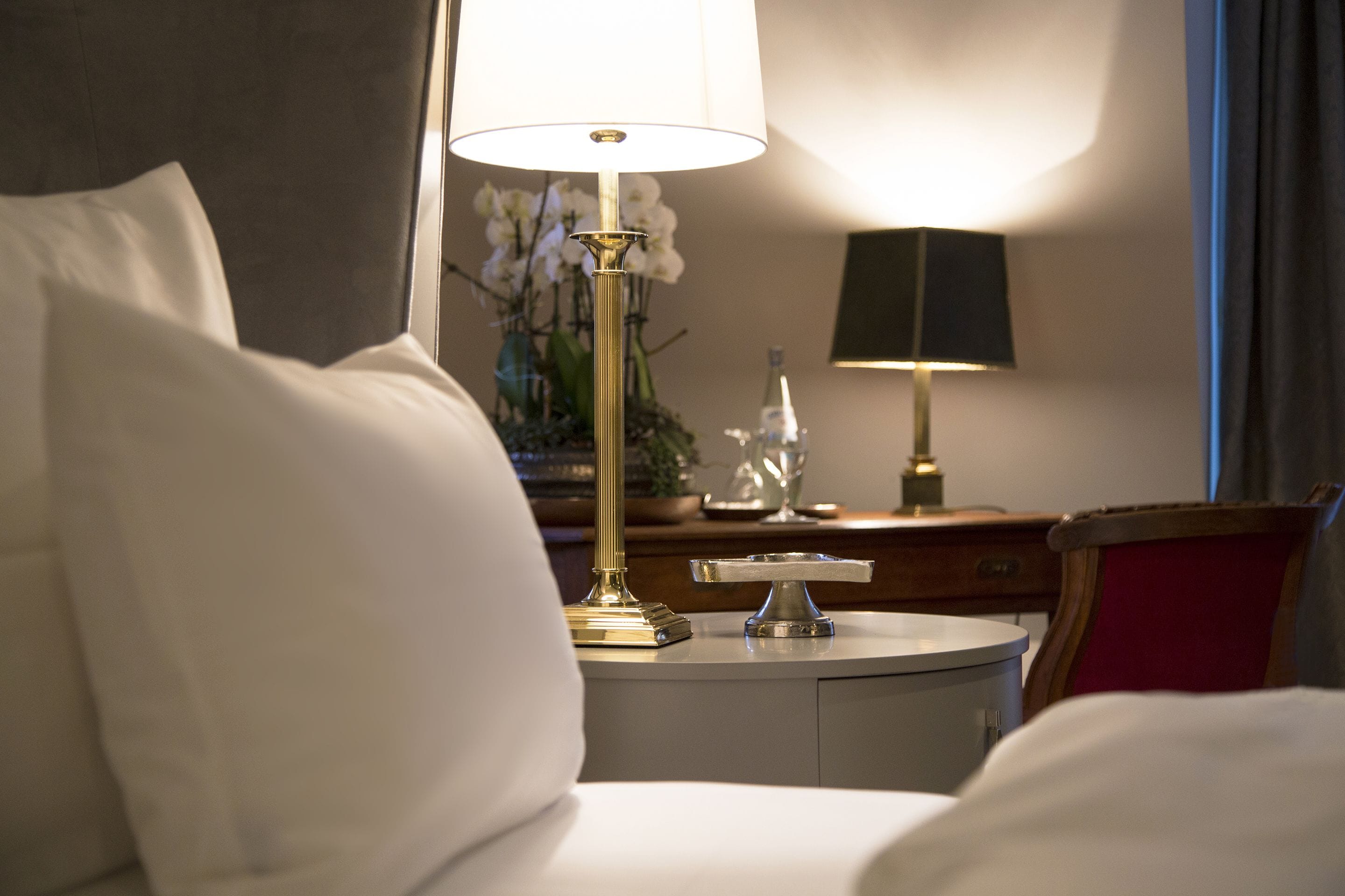 Una habitación de hotel con una cama, una mesita de noche con lámpara, un adorno y un florero con flores.