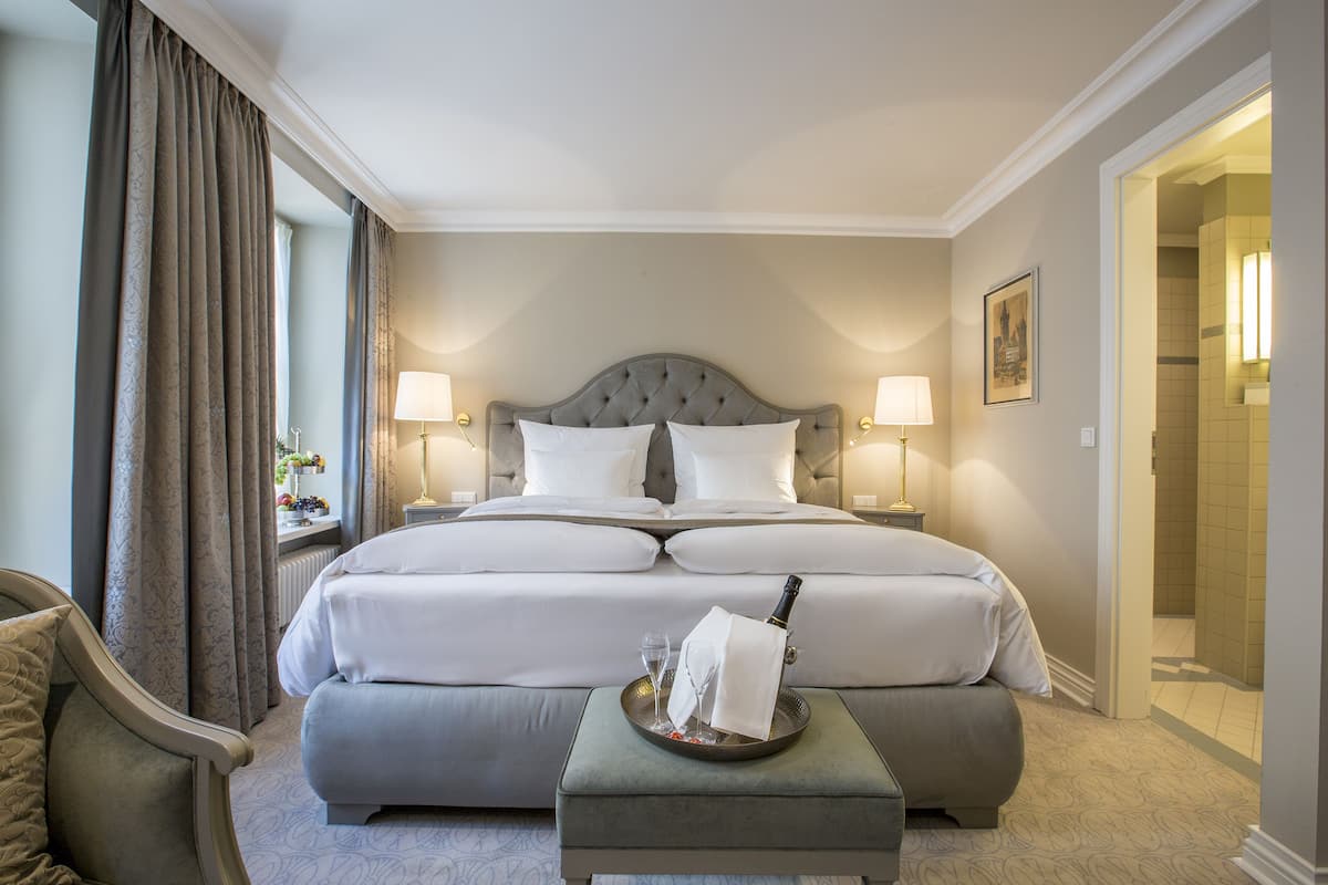 Un lit bien fait, avec du linge de lit blanc, une table de chevet avec une lampe, et un plateau avec une bouteille et des verres posés sur le sol.