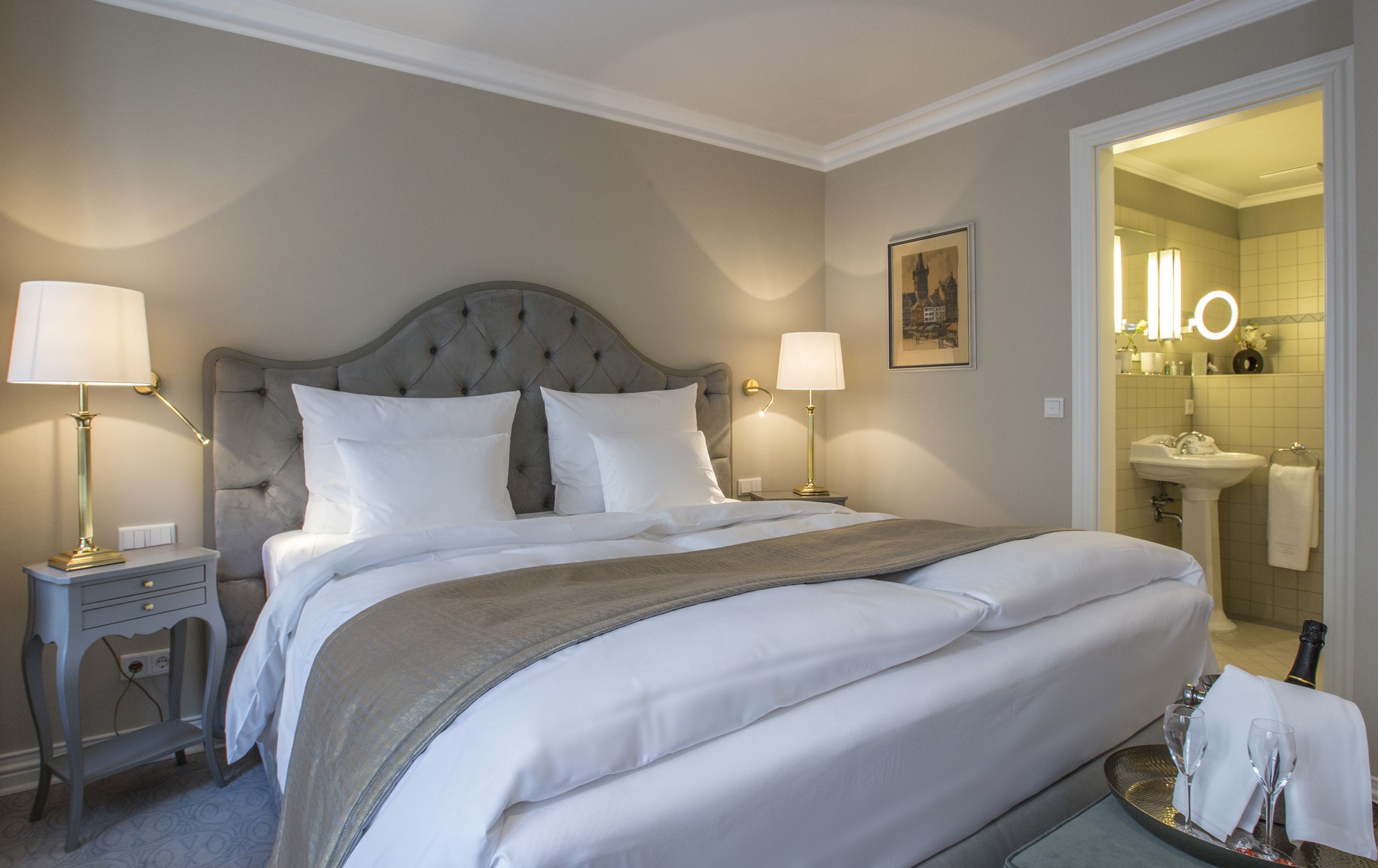A hotel room with a large bed, two bedside tables with lamps, a framed picture on the wall, and a bathroom visible through an open door.
