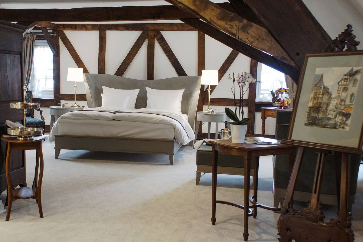 Une chambre à coucher avec un grand lit, des poutres en bois, une petite table avec une lampe et un tableau encadré posé sur un chevalet.