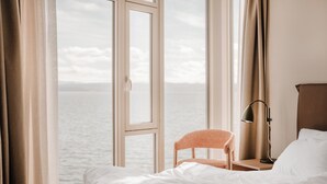 Une chambre d’hôtel avec un lit, une chaise, une table de chevet, une lampe et une vue sur la mer par une fenêtre ouverte.