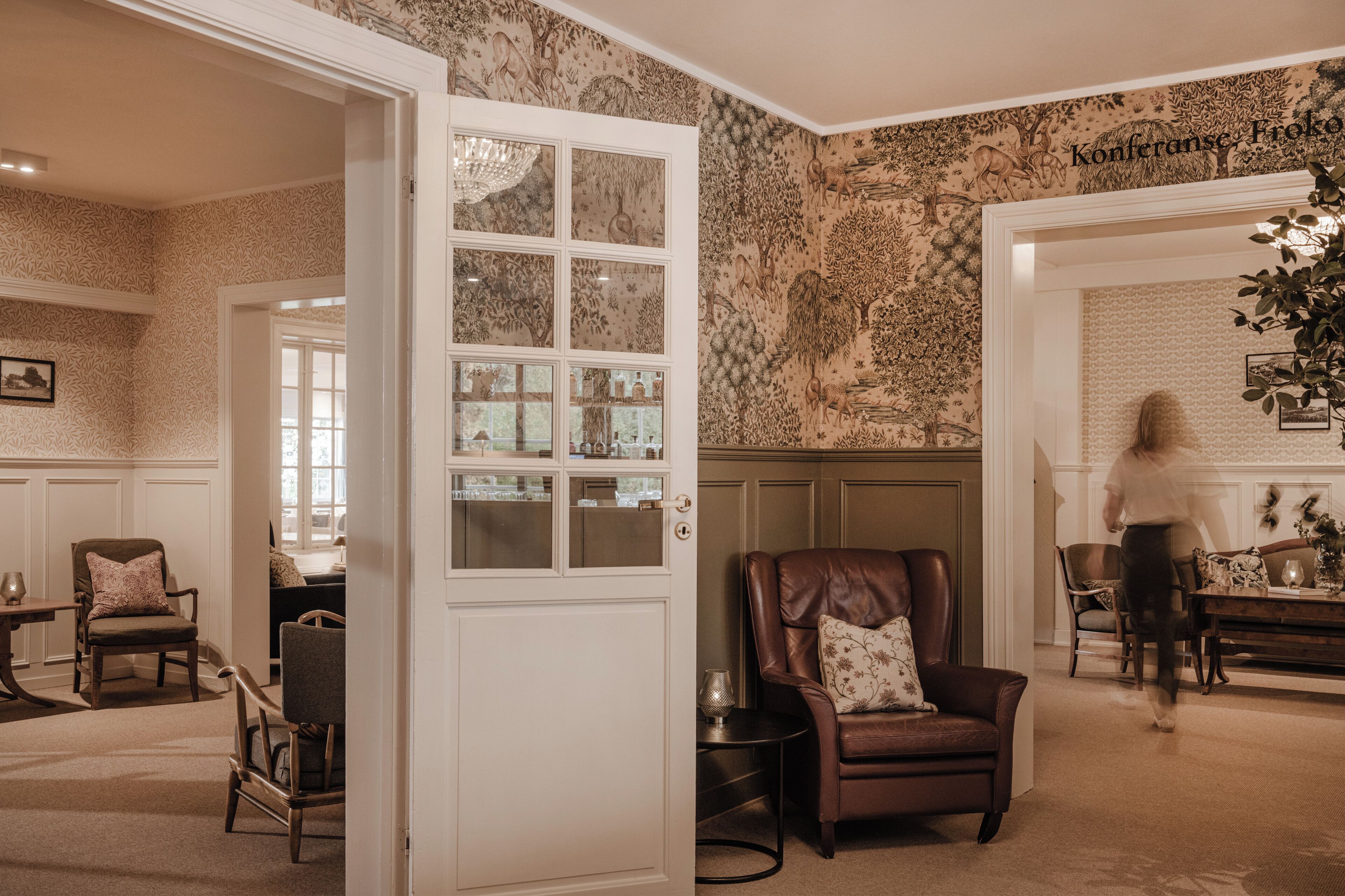 A room with a patterned wallpaper, a white door with glass panels, a brown leather chair, a dining table with chairs, and a chandelier.