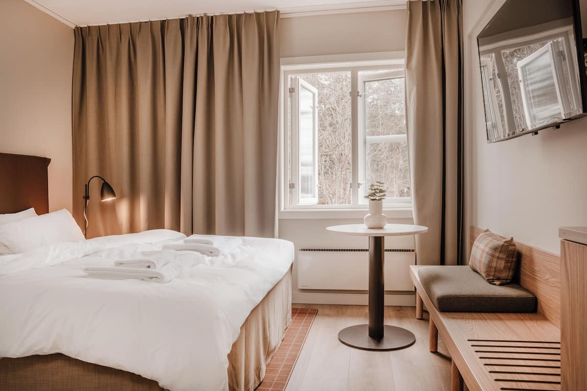 Habitación de hotel con cama, una mesita con una planta, un banco y una ventana con cortinas.