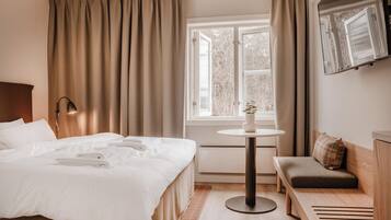 A hotel room with a bed, a small table with a plant, a bench, and a window with curtains.