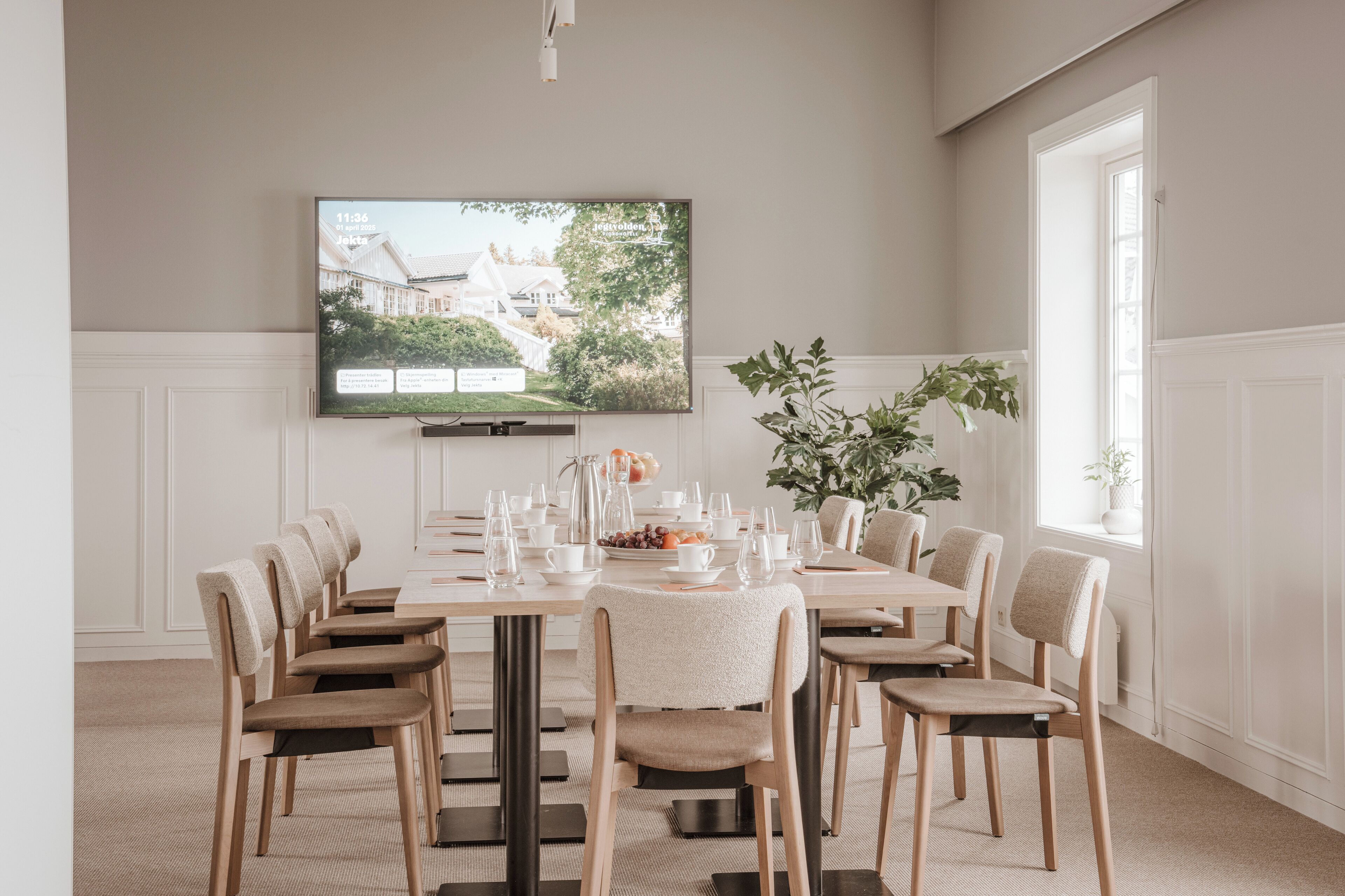 Un comedor con una mesa larga dispuesta para una comida, rodeada de sillas, y un televisor montado en la pared que muestra una vista panorámica.