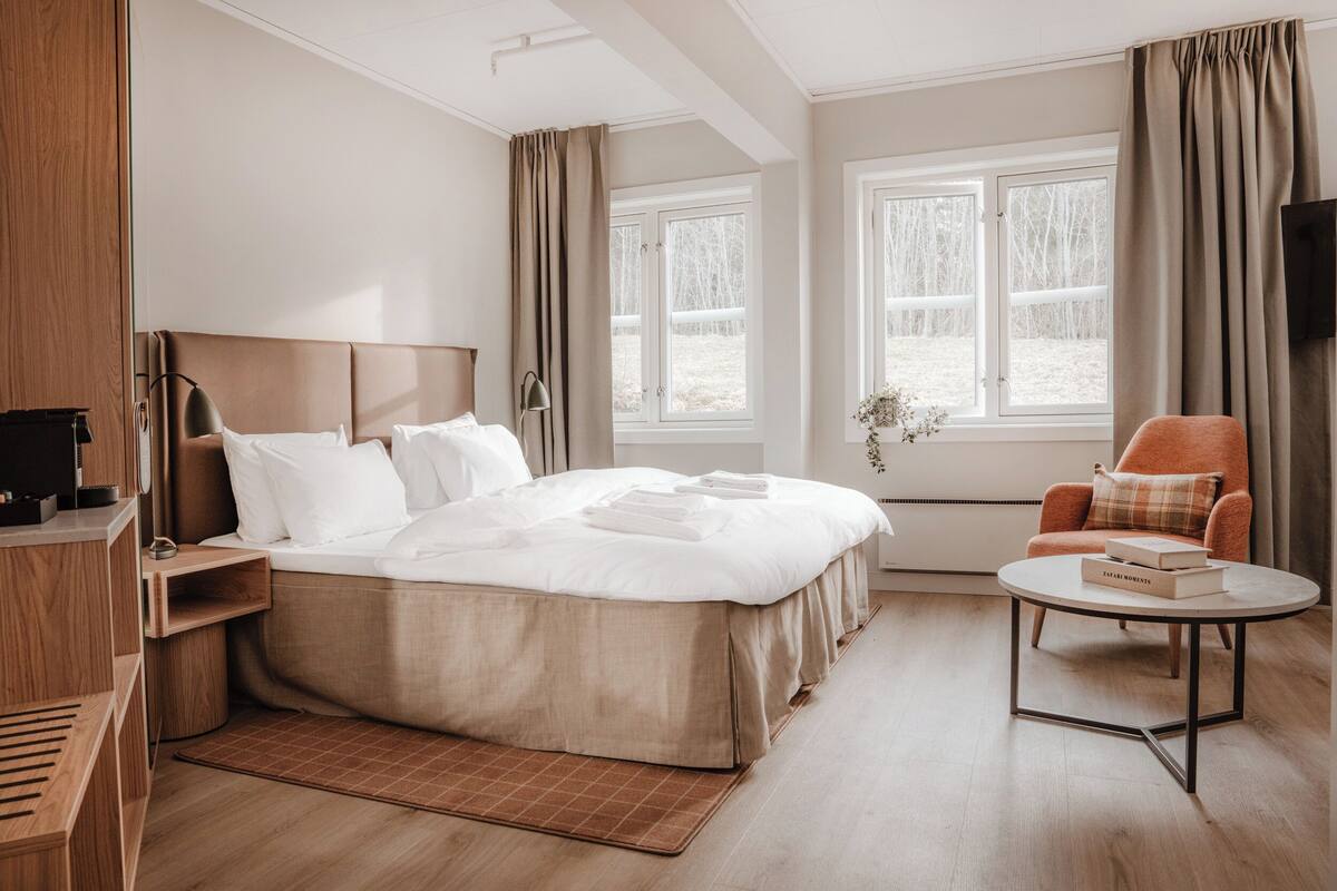 Habitación de hotel con una cama grande, una mesita de noche con lámpara, una silla, una pequeña mesa con un libro y una ventana con cortinas.
