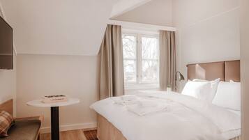 A hotel room with a bed, a small table with a book, a chair, and a window with curtains.