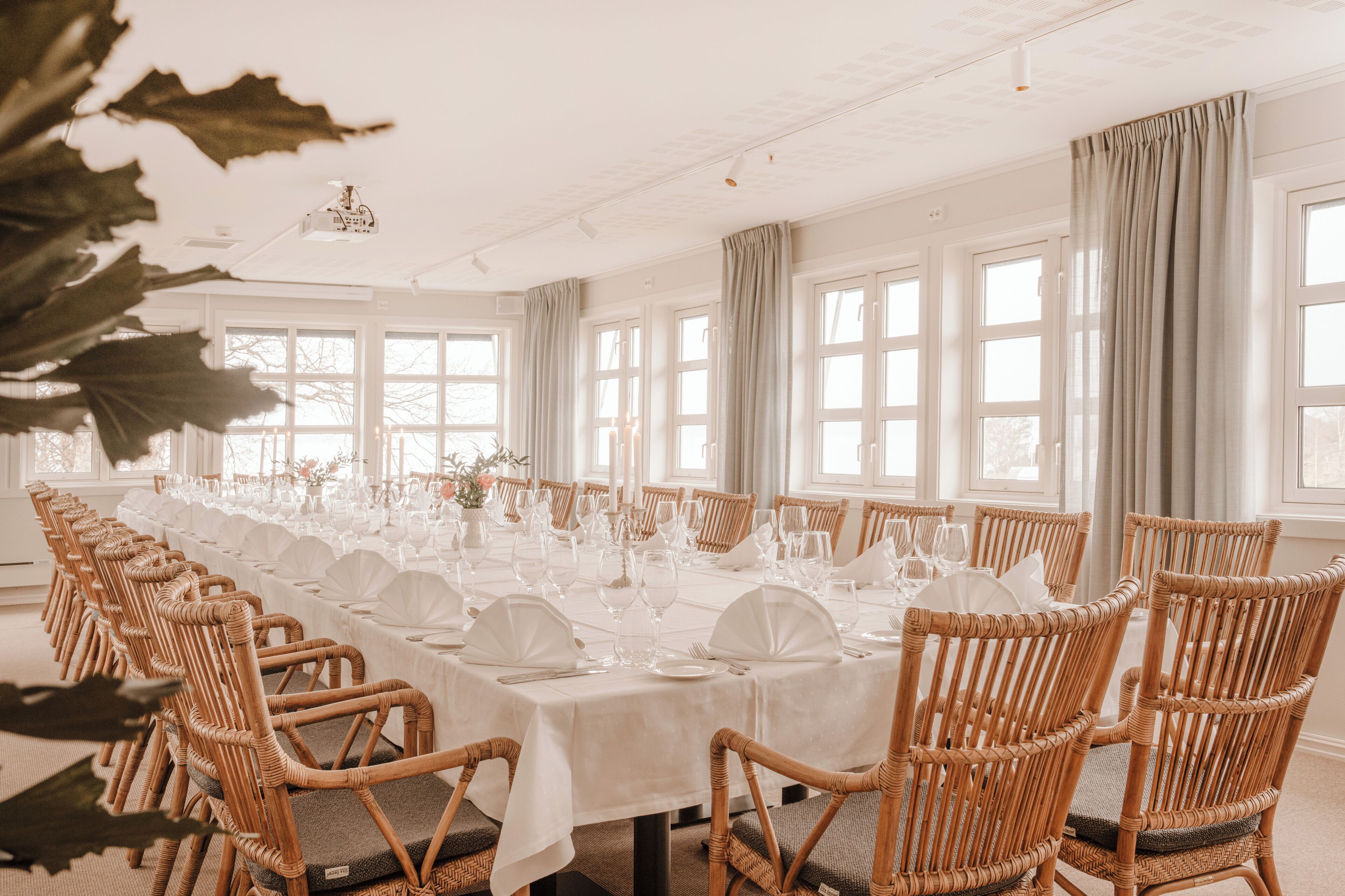 A long dining table set with white tablecloths, wicker chairs, and glassware.