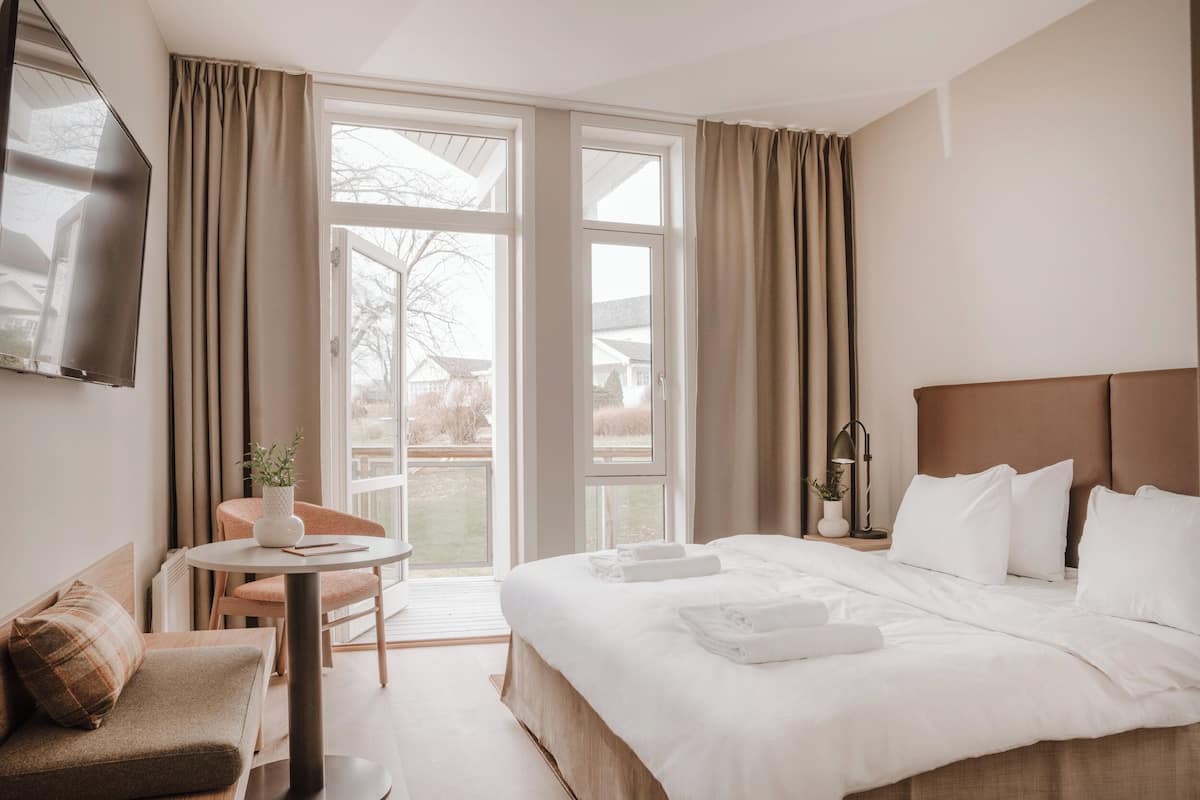 Habitación de hotel con una cama grande, un sofá, una mesita con una planta y vistas al exterior a través de amplios ventanales.