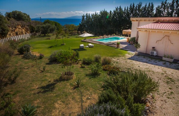 Ein Garten mit Schwimmbad, einer Terrasse mit Liegestühlen und Blick auf das Meer.