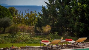 A poolside area with lounge chairs and a table, surrounded by greenery and a view of the sea. - Driftwood Villa (Kefalonia)