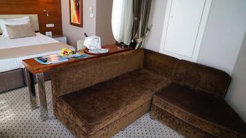 Ein Hotelzimmer mit einem Bett, einem braunen Sofa, einem Holztisch mit einer Zeitschrift, einer Obstschale und einem Vorhang.