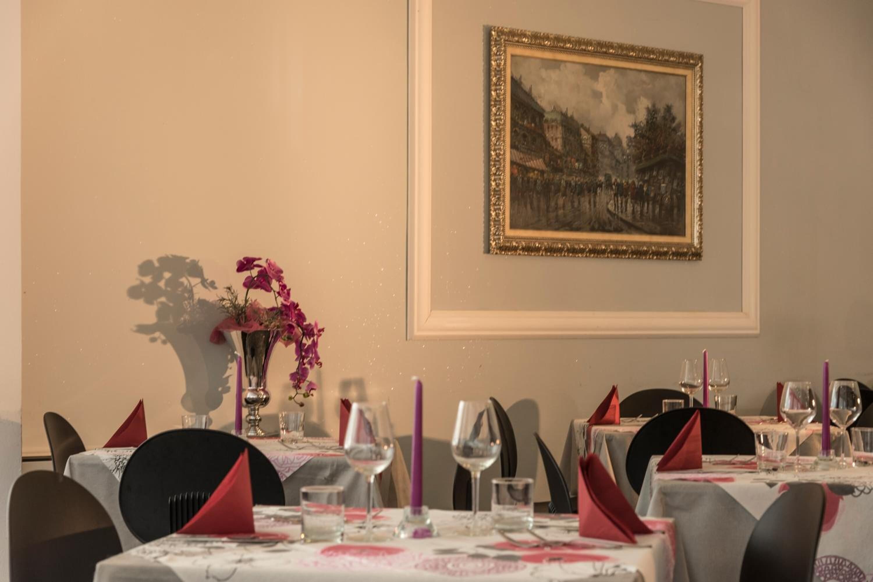 A dining room with tables set for a meal, a framed painting on the wall, and a vase with flowers on the table.