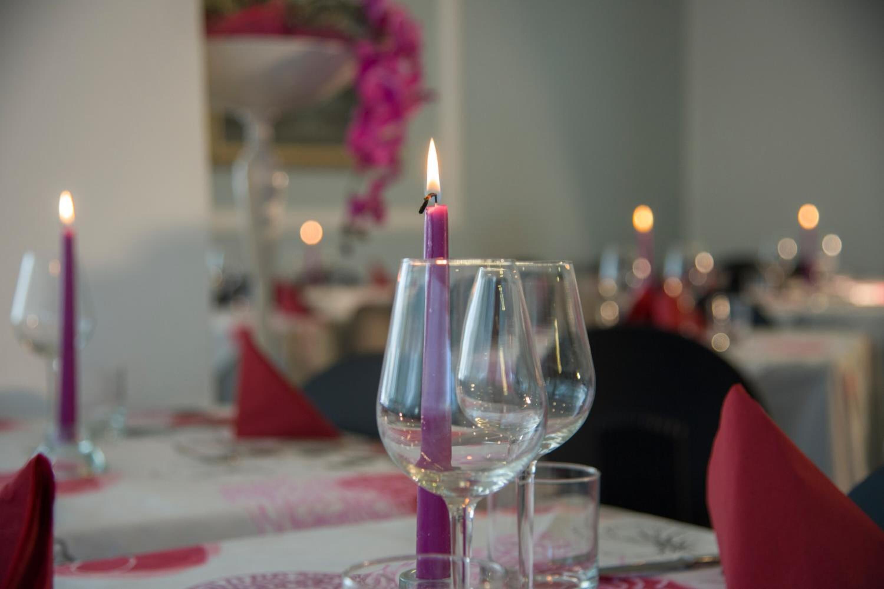 A dining table with a candle in a glass, surrounded by empty wine glasses and red napkins.