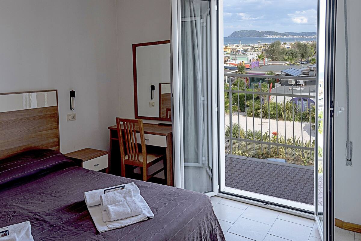 A hotel room with a bed, a desk, a chair, and a large window with a view of the city and mountains.