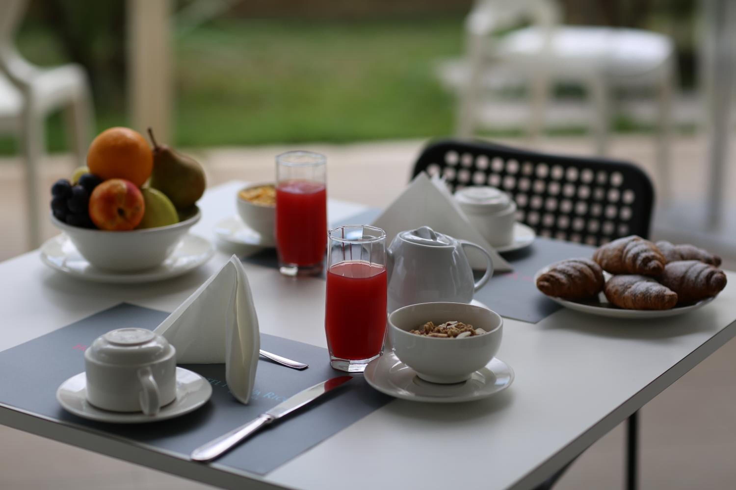 A breakfast table with fruits, a glass of red juice, a bowl of cereal, a coffee pot, and croissants.