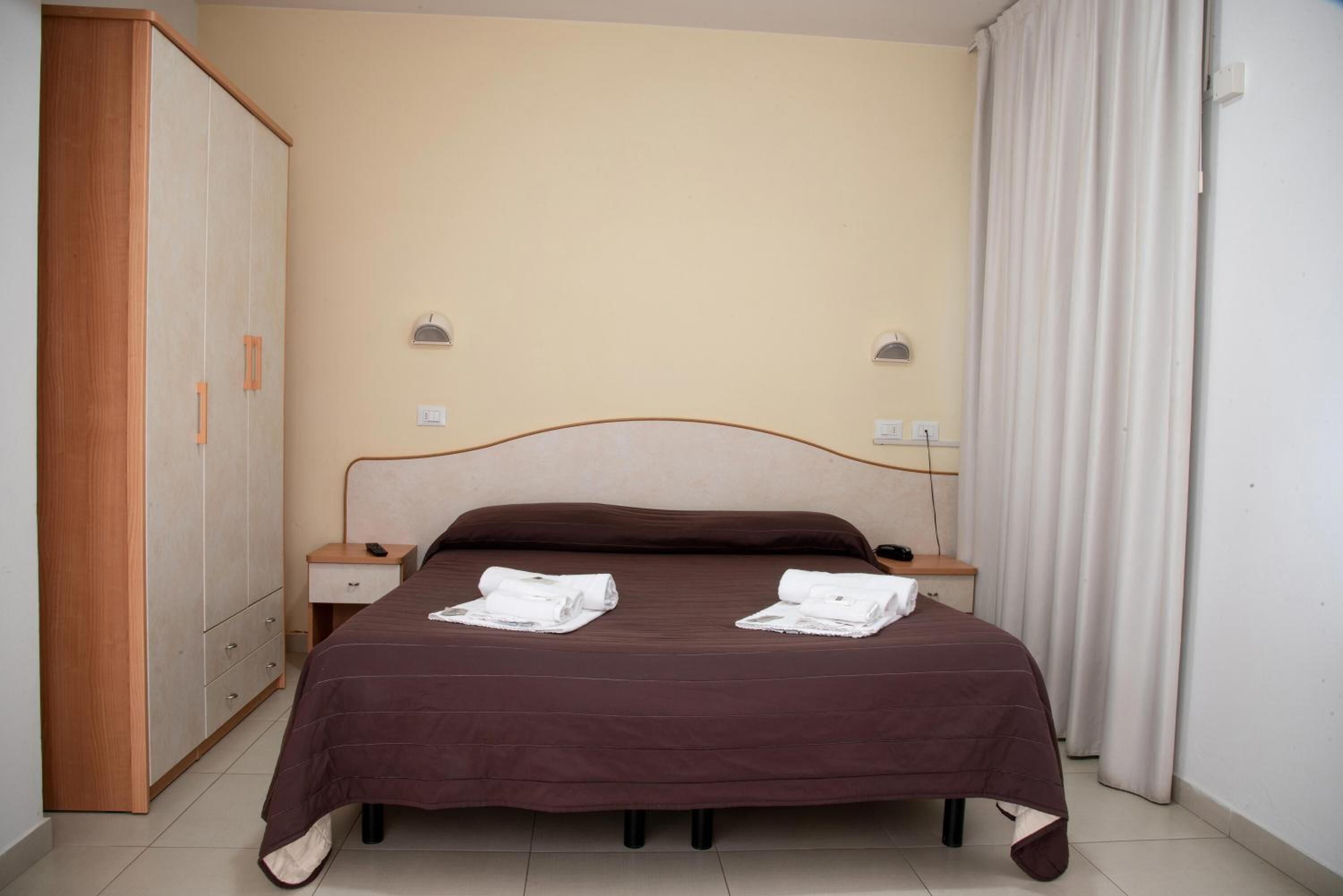 A neatly made bed with a brown bedspread and two rolled towels, a wooden wardrobe, and a nightstand with a lamp.