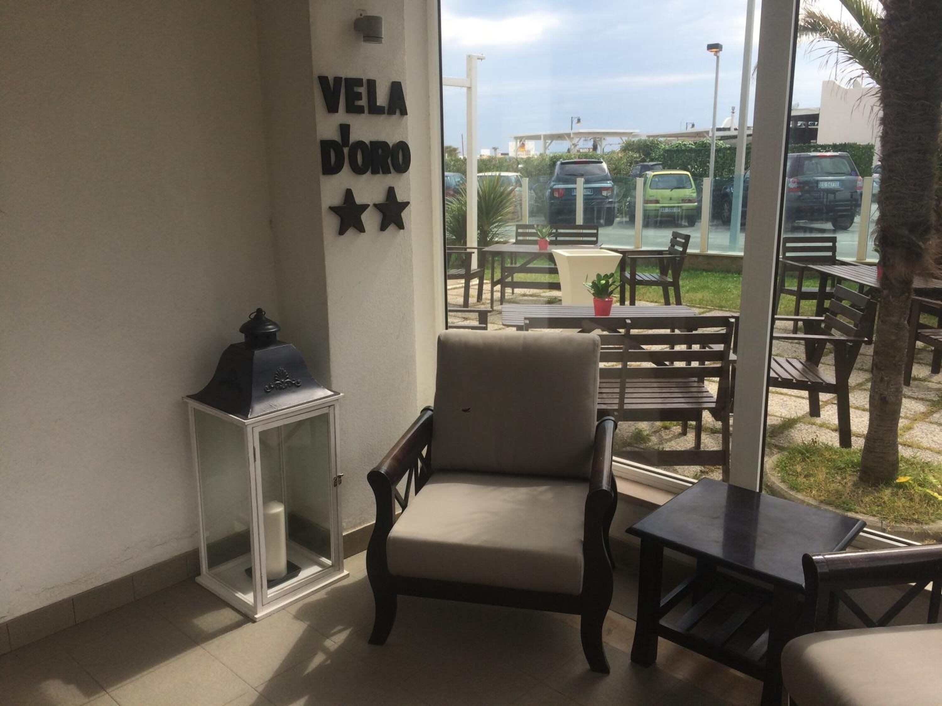 A room with a seating area, a wall-mounted sign reading "Vela D'oro", and a view of parked cars outside.