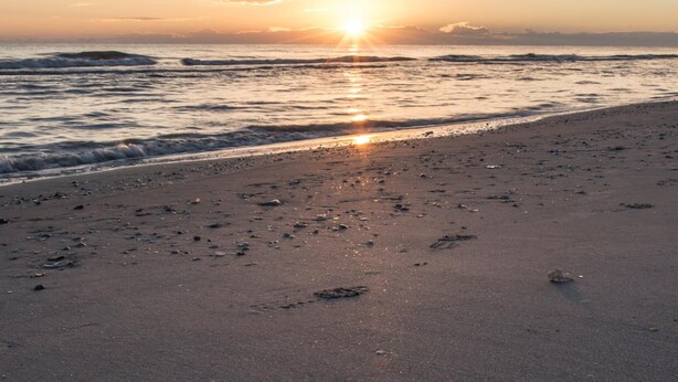 Een strand bij zonsondergang, met de zon laag aan de horizon, golven die zachtjes op het strand komen en verspreid afval op het zand.