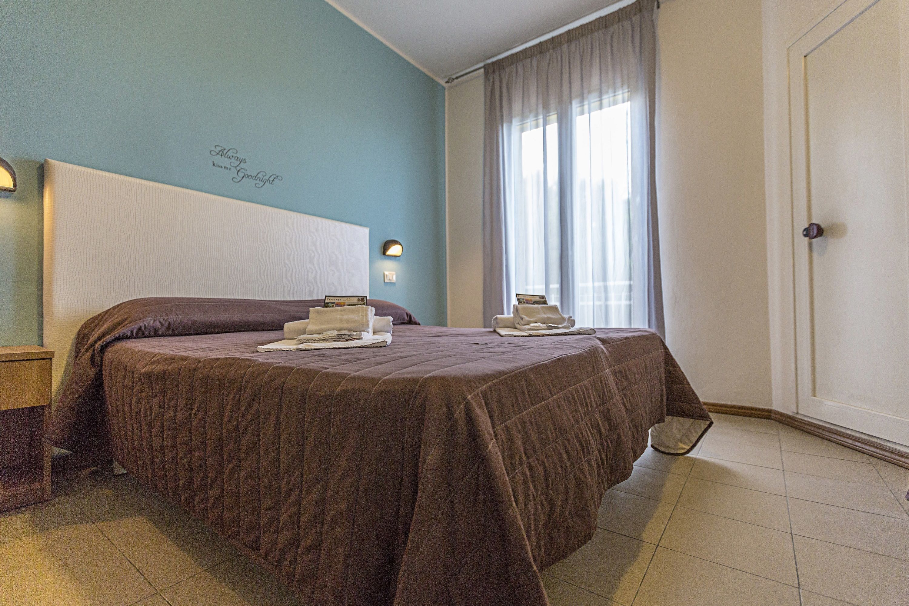 Una camera d'albergo con un letto, un comodino, una sveglia a muro e una finestra con tende.
