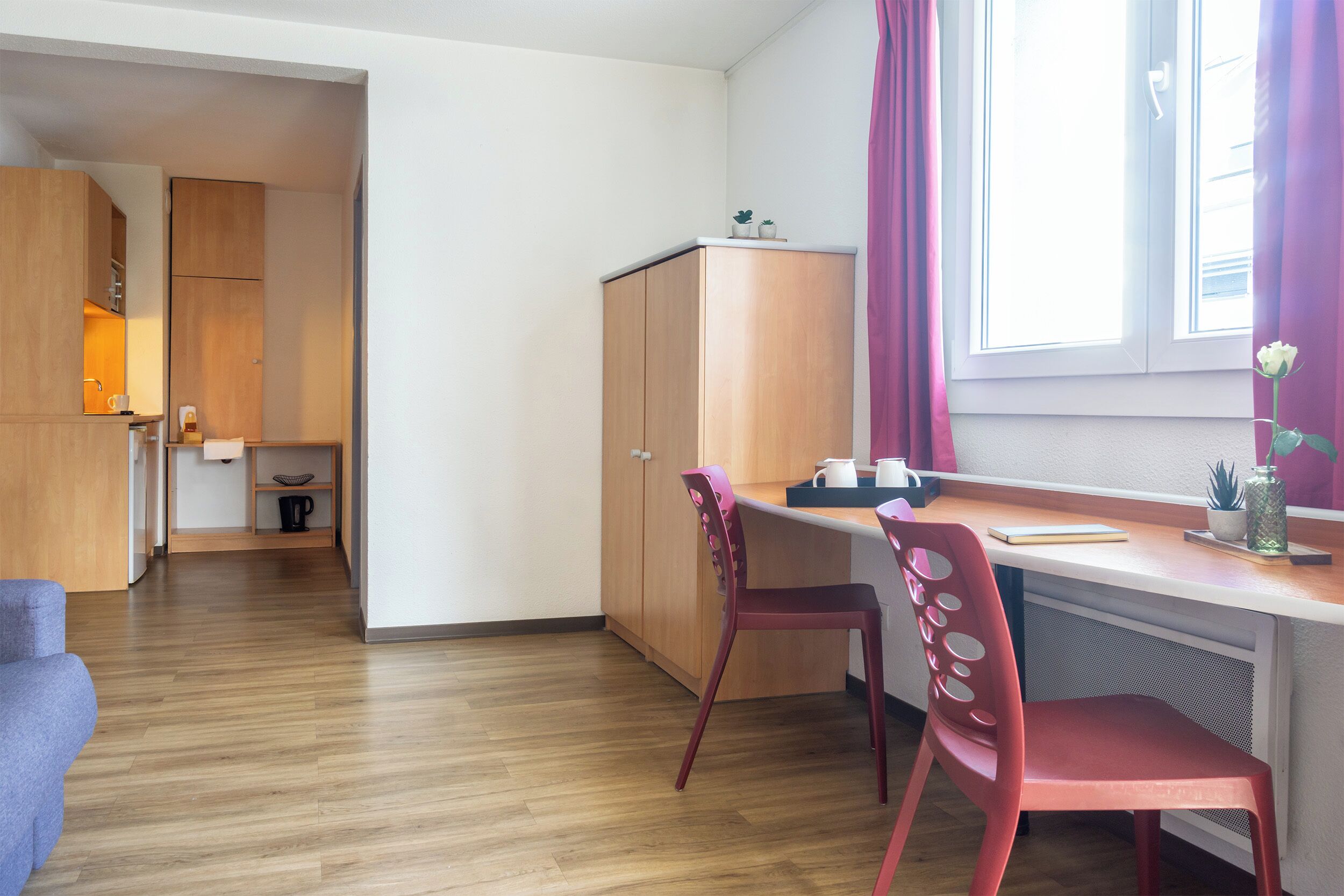A compact kitchen with wooden cabinets, a dining area with a red chair, and a window with curtains.