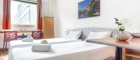 Habitación de hotel con dos camas, un escritorio, sillas, un florero con flores y un cuadro en la pared.