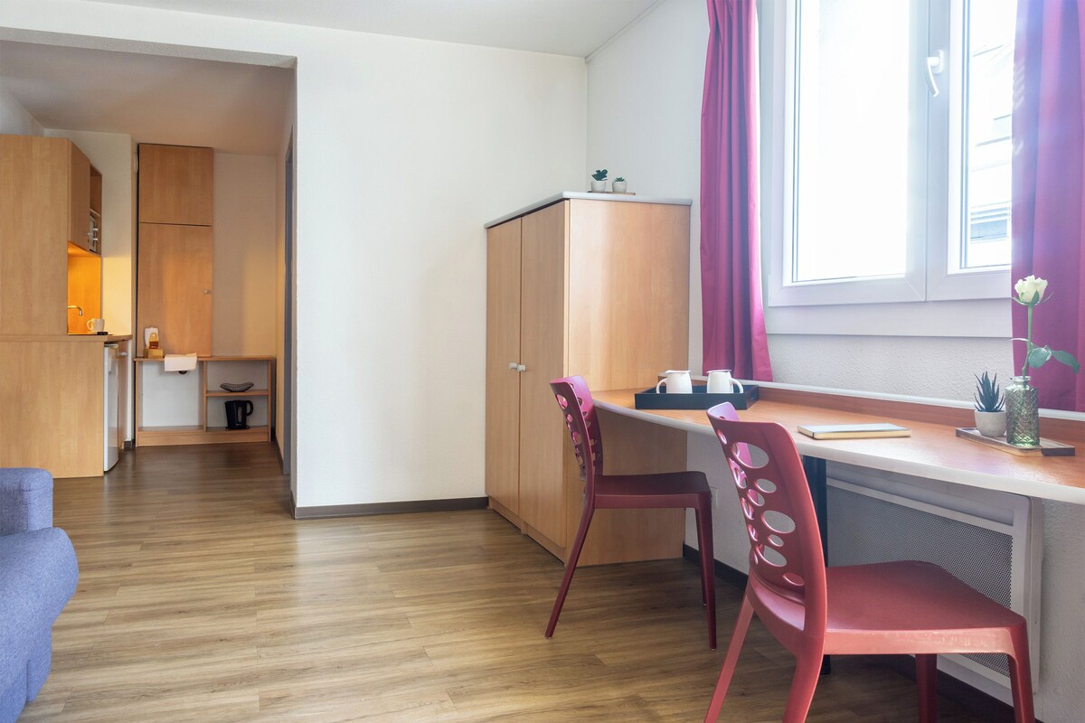 Una cocina compacta con armarios de madera, un comedor con una silla roja y una ventana con cortinas.